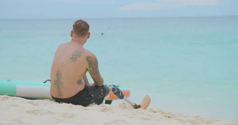 Tired surfer with bionic leg sitting on sandy beach and relaxing after surfing. Video stock 187698383