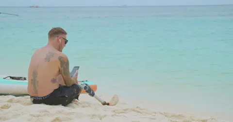 Tired surfer with bionic leg sitting on sandy beach and relaxing after surfing. Stock Footage 187699494