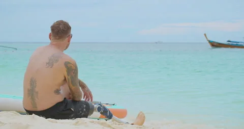 Tired surfer with bionic leg sitting on sandy beach and relaxing after surfing. Stock Footage 242443693