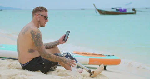 Tired surfer with bionic leg sitting on sandy beach and relaxing after surfing. Video stock 242443803