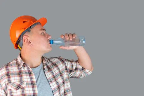 A tired worker drinks water from a plastic bottle standing on a gray backgrou Stock Photos