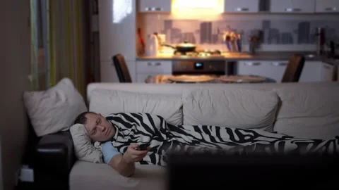 Tired young man lying on a sofa covered with a plaid, watching TV Stock Footage 131729634