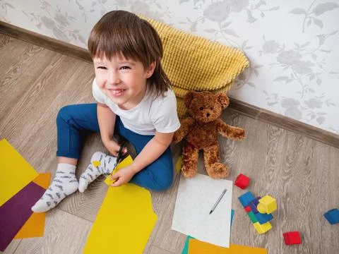 Toddler boy learns to cut colored paper with scissors. Kid sits on floor in k 库存照片