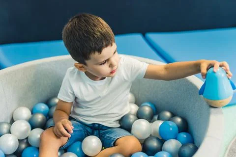 Toddler boy playing with a toy while sitting in a ball pit full of colorful Stock Photos