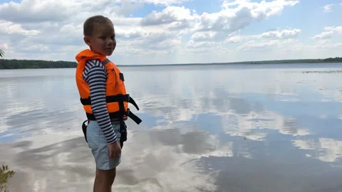 A toddler in a bright orange life jacket stands by the calm water, highlighting  Stock Footage 340508089