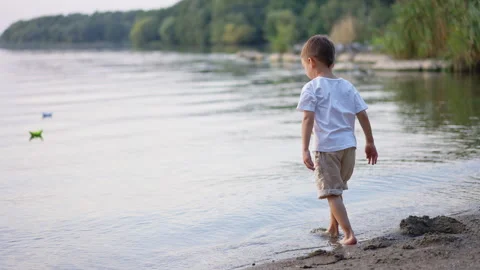 Toddler crouching at river shore touching water on sandy bank. Side view with Stock Footage 336382439