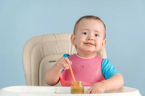 Toddler eats broccoli vegetable puree with an orange plastic spoon while sitt Stock Photos