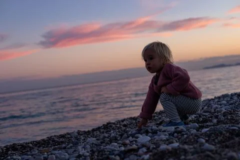 A toddler exploring pebbles on a beach during sunset with colorful clouds Stock Photos