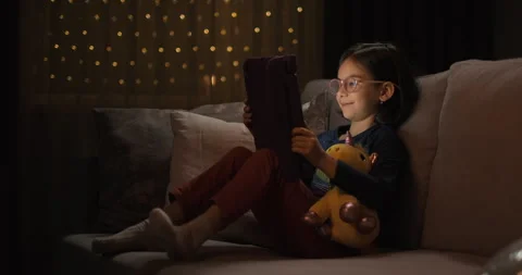 Toddler girl watching at the tablet while sitting on sofa at night Stock Footage 243077374
