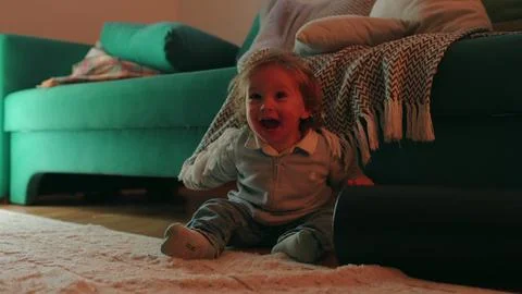 Toddler hiding beneath patterned blanket beside living room sofa at home, e.. Stock Photos