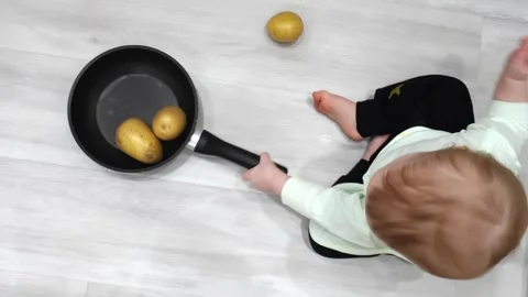 Toddler joyfully plays on the floor with a kitchen skillet and potato Stock Footage 266751623