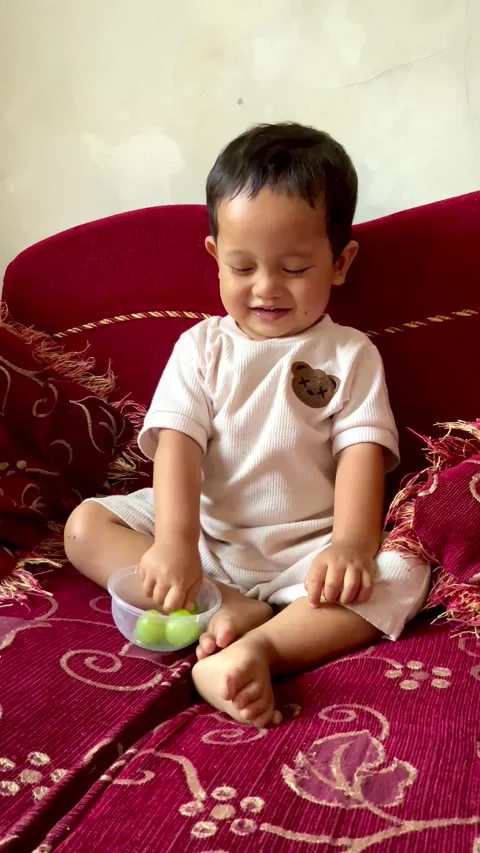 Toddler playing with small green grape on a red sofa Stock Footage 344104476