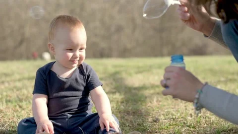 A toddler sitting on the grass smiles while watching bubbles being blown by an Видео 278073230