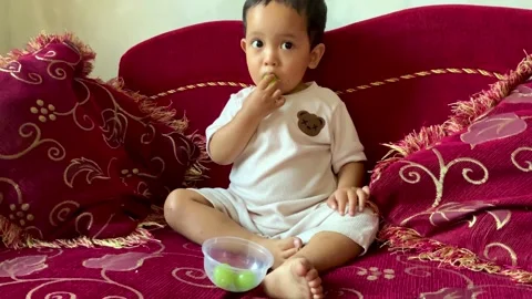 Toddler sitting on a red sofa eating green grapes Stock Footage 344104495