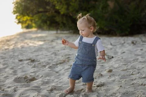 Toddler in striped overalls exploring a sandy beach at sunset Stock Photos