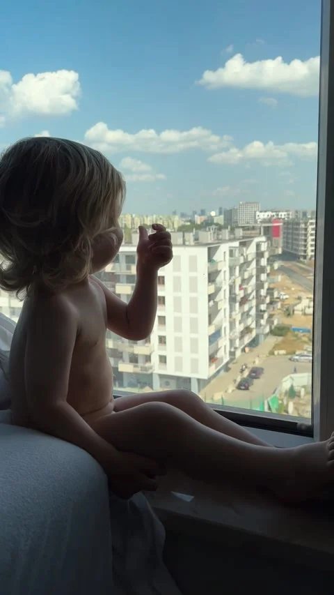 Toddler on windowsill reaching toward window glass while gazing at urban street Stock Footage 345499798