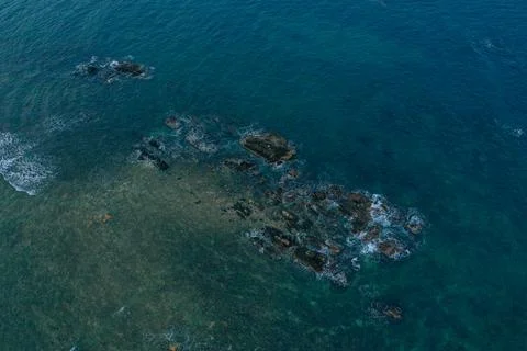 Top-down aerial view of dark coastal rocks breaking the surface of deep blue Stock Photos