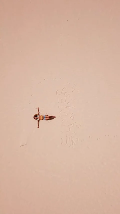 Top-down aerial view of a young woman in a swimsuit sunbathing on a vast pink 스톡 동영상 344839513