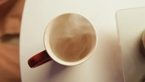Top-down hand lifting steaming coffee cup beside laptop corner on light desk Stock Footage 343542167