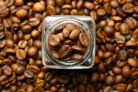 A top-down view of roasted coffee beans scattered around a square glass jar.. Stock Photos