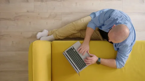 Top down view of sofa next to which man sits down on floor with laptop and work Stock Footage 243607708