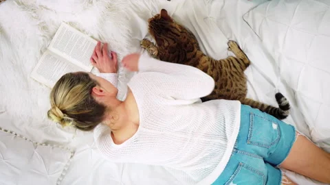 Top-down view of a woman lying on the bed reading a book with Bengal cat nearby Stock Footage 150840026