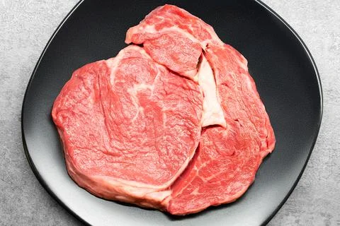 Top studio shot of a raw beef rib eye steak on a black design plate standing on Stock Photos