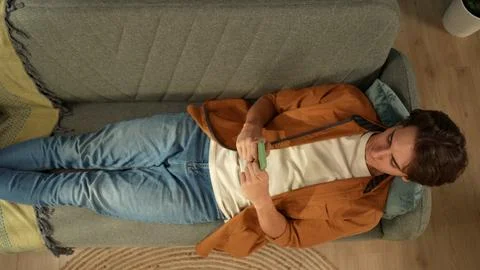 Top view capturing a young man laying on the couch, looking at the screen of his Stock Photos