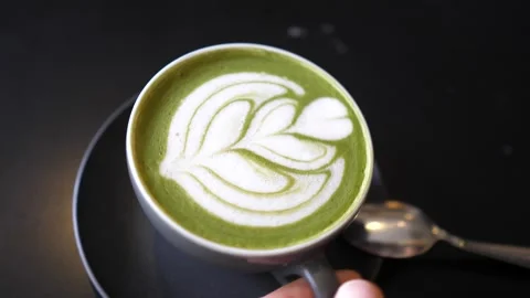 Top view of a hand picking up black cup of matcha tea latte with latte art on it Stock Footage 148992032