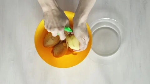 Top view of hands peeling fresh potatoes with a green peeler over an orange.. Stock Footage 315973633
