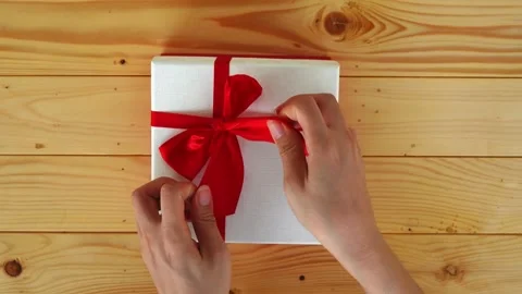 Top View Hands Unwrapping White Gift Box with Red Ribbon Bow on Wooden Surface Stock Footage 310921552