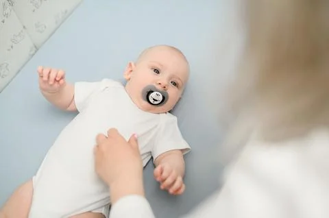 Top view of infant with pacifier lying in crib and mom bending over baby in Stock Photos