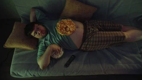 Top view of lazy man lying on sofa with pillows watching TV and eating popcorn Stock Footage 314396651