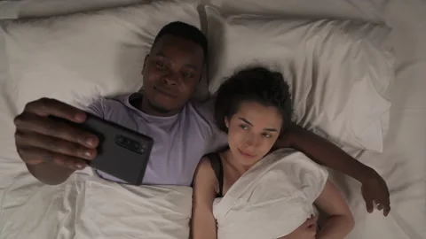 Top view of mixed-race couple lying in bed at night and taking selfie with Stock Footage 136581706