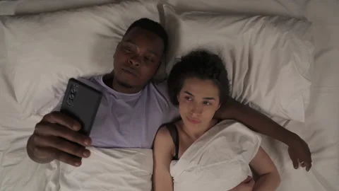 Top view of multi-ethnic couple lying in bed at night and talking with friends Stock Footage 136582074