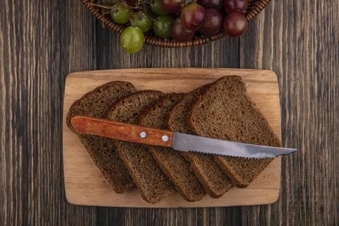 Top view of rye bread slices and knife on cutting board with basket of grap.. Фото