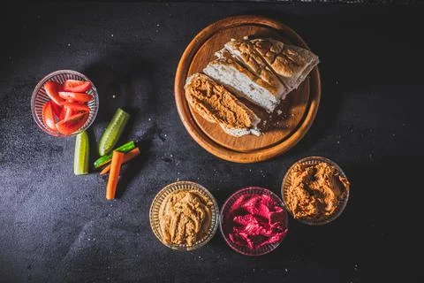 A top view of sliced bread, vegetables and bowls of homemade hummus on a blac Stock Photos