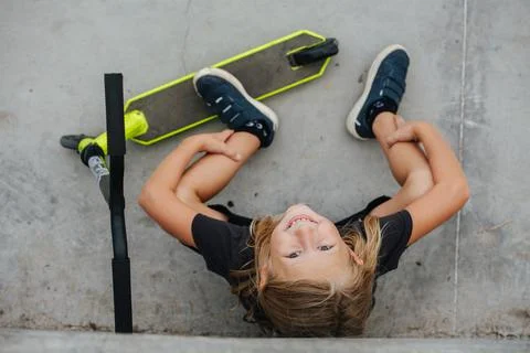 Top view of a smiling boy sitting on concrete floor next to a scooter Stock Photos