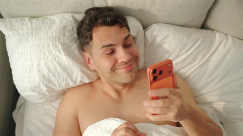 Top view of smiling man in bed texting on orange phone and grinning. Vídeo Stock 348192488