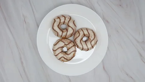 Top View Spinning Donuts Covered In White Frosting With Brown Swirls On A White  스톡 동영상 150066408