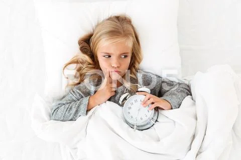 Top view of thoughtful little girl lying in bed with alarm clock, looking asi Stock Photos
