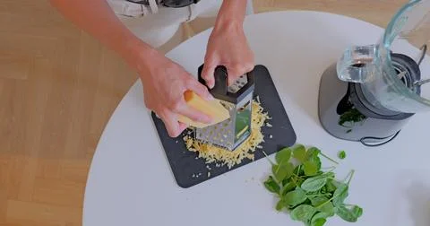 Top View of Woman Grating Cheese with Spinach and Blender Nearby in Kitchen 库存照片