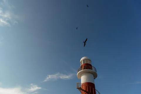 Top of a white-red lighthouse against a blue sky with an airplane. Stock Photos