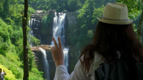 Tourist gesturing peace sign while admiring waterfall in green valley Stock Footage 311609423
