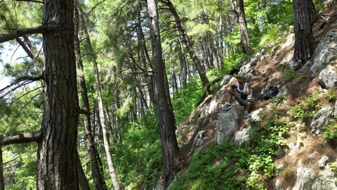 A tourist guy is sitting on a mountainside in a pine forest among rocks Stock Footage 249474085