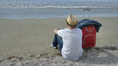 Tourist in a hat sits on the beach and looks at the waves Stock Footage 123732757