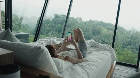 Tourist using smartphone on bed in modern hotel room with forest view Stock Footage 305356095