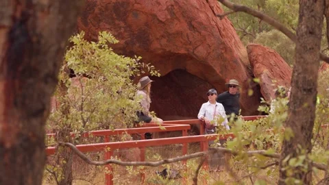 Tourists Pausing On Uluru Base Walk Boardwalk Beside Rock Cave Framed By Native Stock Footage 336113111