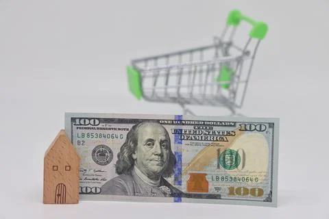Toy house with a banknote on the background of an empty toy grocery cart, ris Stock Photos