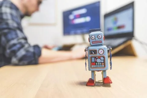 Toy robot on a desktop, man working on the computer in the background. Symbol 스톡 사진